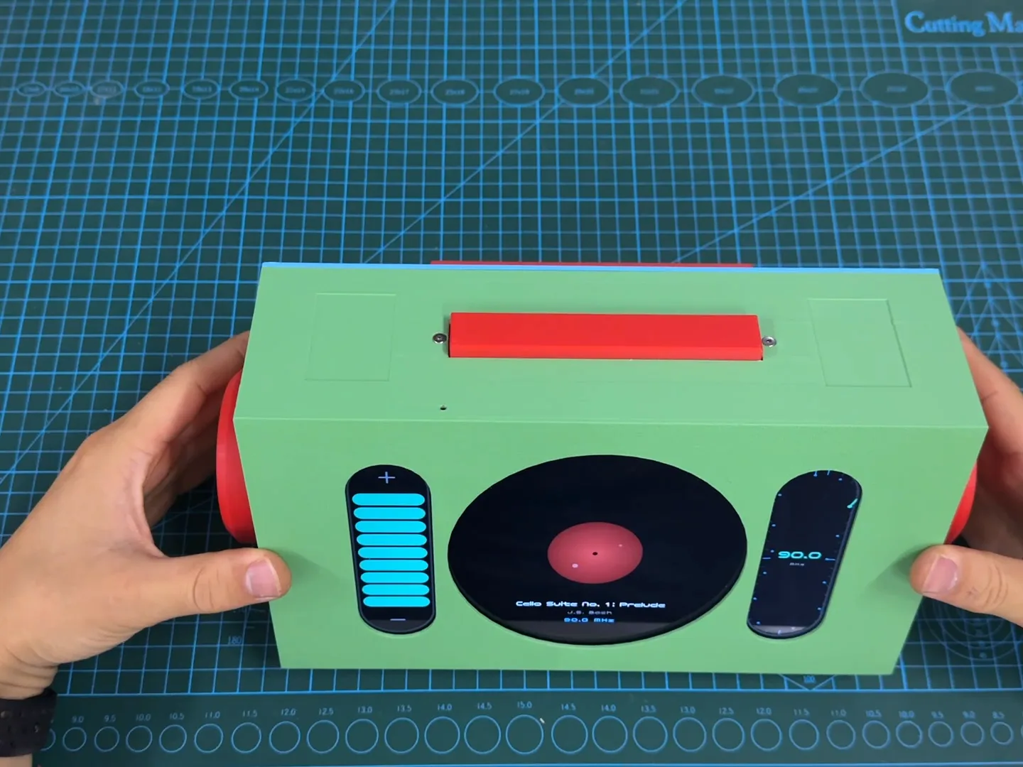 Two hands holding a green 3D-printed radio with red knobs on both sides, a red button on top and a screen showing a vinyl record