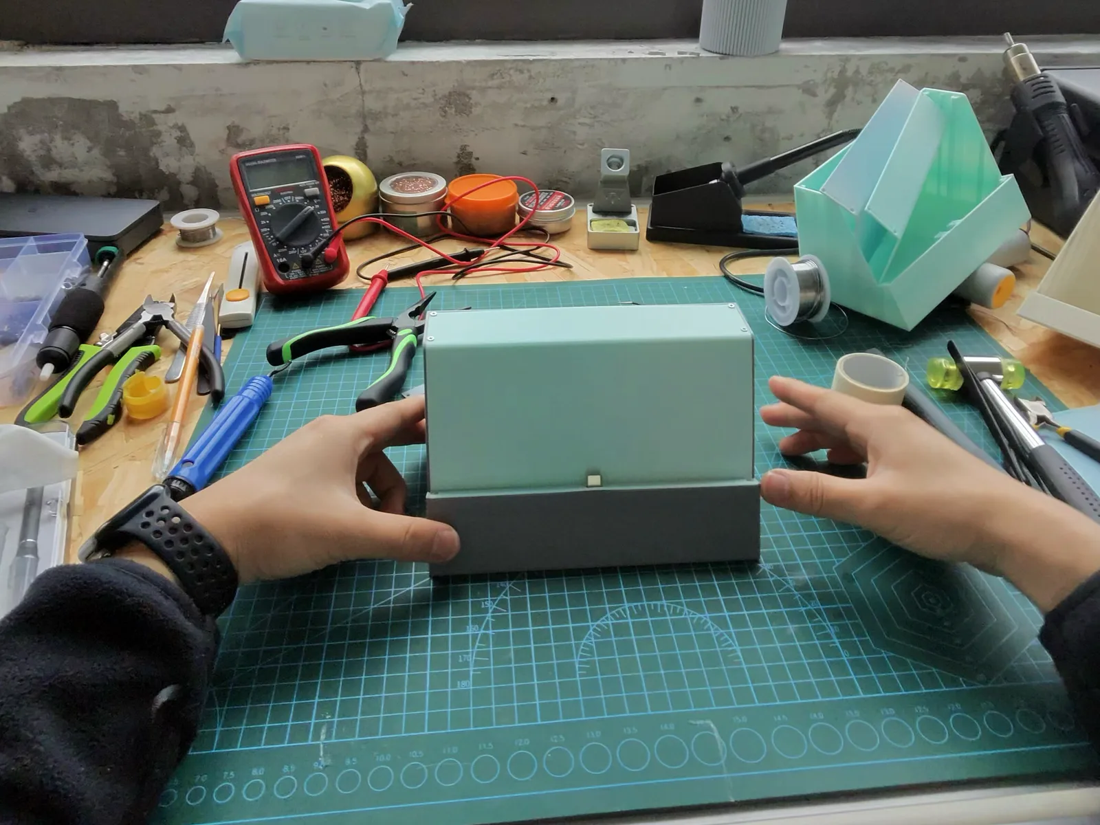 Two hands fitting a mint-green printed enclosure together on a cutting mat at a workbench under a window