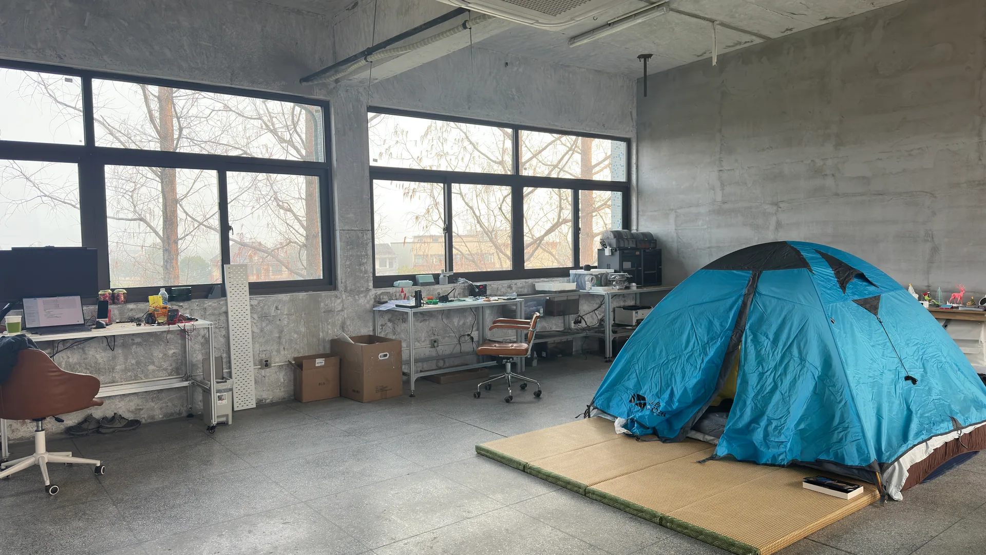 A concrete classroom with workbenches along a row of windows and a blue tent on the floor