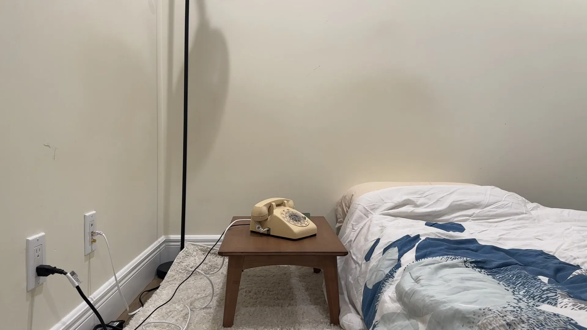 A cream rotary telephone on a low wooden table beside a bed