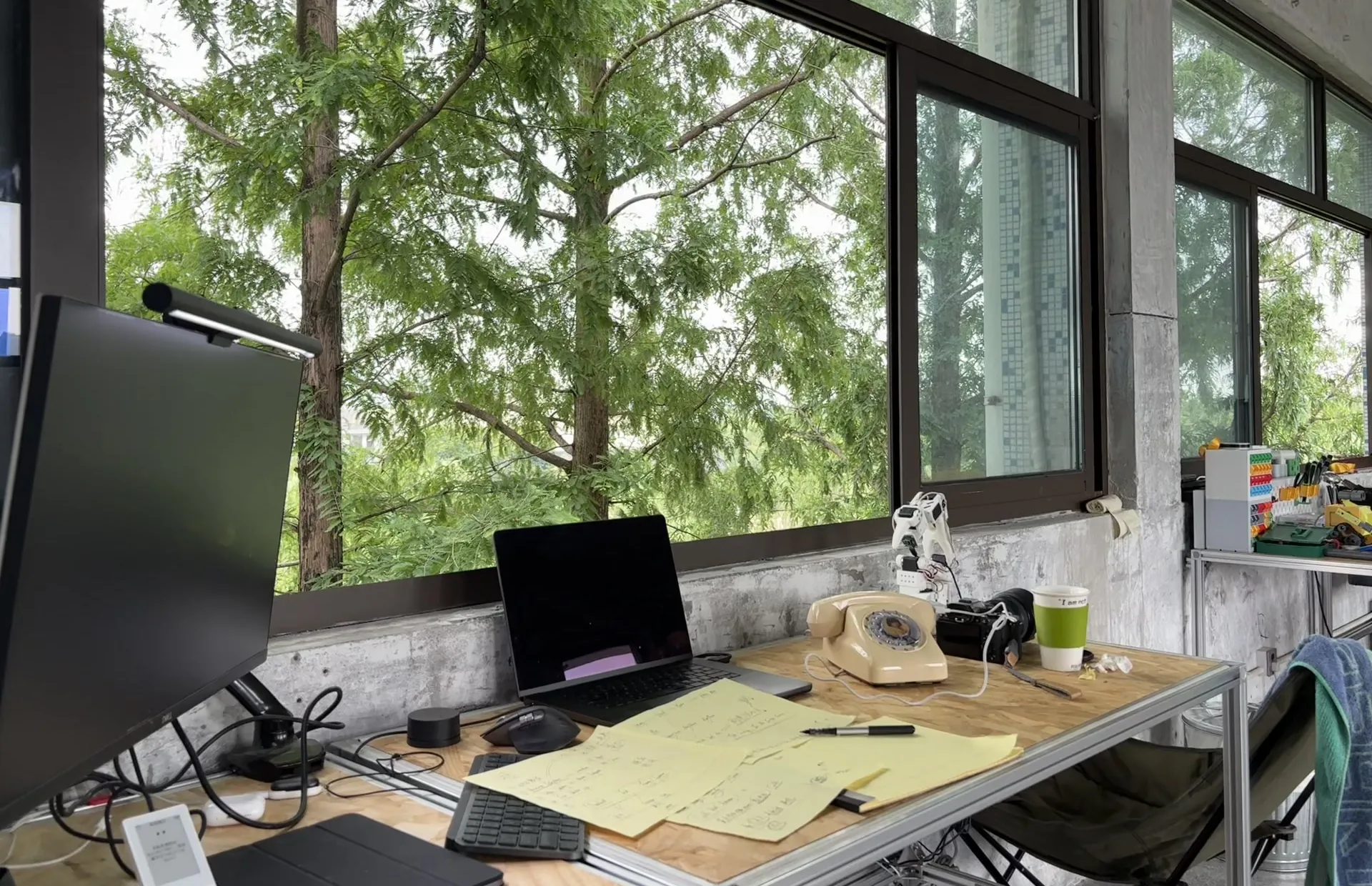 A plywood desk with a laptop, notes and a rotary telephone under tall windows full of trees